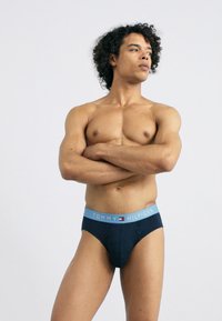 Námorné bavlnené slipy s svetlomodrým elastickým pásom s logom "TOMMY HILFIGER". Hladká textúra; priliehajúci dizajn. Model pózuje s prekríženými rukami.