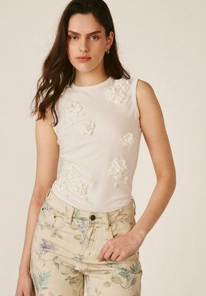 Joven mujer con cabello largo y oscuro lleva una camiseta blanca sin mangas con aplicaciones florales en 3D y vaqueros claros de talle alto con estampado floral.