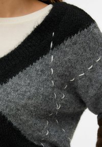 Oprta pletena majica z vzorcem krpanke v sivi, črni in beli barvi. Teksturirana z ohlapnimi šivi in okroglim izrezom. Mehki, puhasti material.