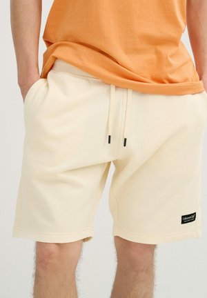 Persona que lleva pantalones cortos color crema con cordón y una camiseta naranja, manos en los bolsillos, de pie frente a un fondo liso.