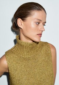 Maglione lavorato a maglia in giallo senape con colletto alto a coste, caratterizzato da un filato testurizzato e un design senza maniche. Finitura in tessuto liscia.