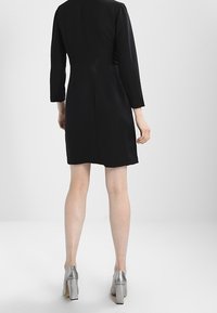 Vestito nero all'altezza del ginocchio con maniche a tre quarti, silhouette aderente, tessuto liscio e cucitura posteriore. Abbinato a tacchi alti metallici.