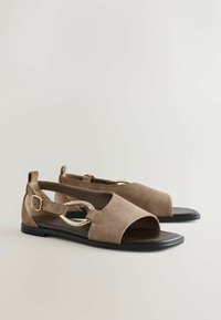 Paire de sandales plates en daim beige à bout ouvert, semelles noires, brides de cheville avec boucles dorées et grands accents circulaires dorés sur les côtés.