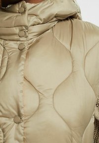 Jasnotrwa beżowa pikowana kurtka puchowa z wysokim kołnierzem, zapinana na guziki, o gładkiej, błyszczącej teksturze tkaniny.