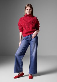 Roter gestrickter Pullover mit Rippdesign, hohem Kragen und halbem Reißverschluss. Kombiniert mit weit geschnittenen blauen Jeans und roten Ballerinas.