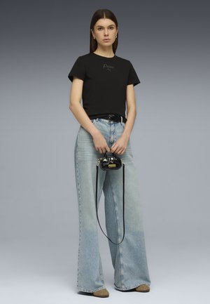 Donna che indossa una t-shirt nera Puma e jeans azzurri a gamba larga, con in mano una piccola borsa nera Puma con tracolla lunga.