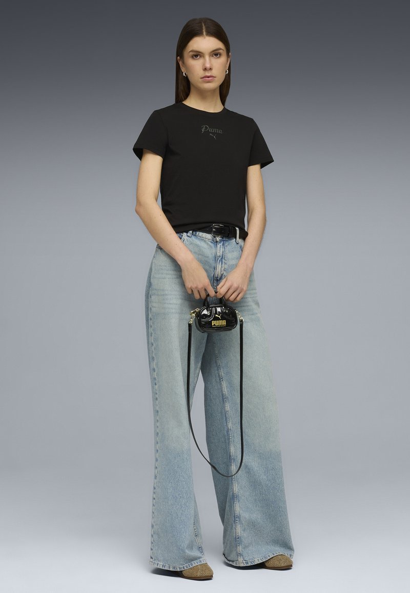 Femme portant un t-shirt noir Puma et un jean large bleu clair, tenant un petit sac à main noir Puma avec une longue bandoulière.