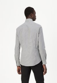 Mann steht mit dem Rücken zur Kamera, trägt ein schwarz-weiß gestreiftes Langarmhemd und schwarze Hosen vor einem weißen Hintergrund.