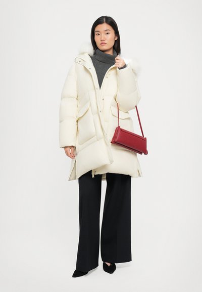 Weißer Daunenmantel mit einer mit Fell gefütterten Kapuze, zwei großen vorderen Taschen und einem durchgehenden Reißverschluss. Kombiniert mit einem grauen Rollkragenpullover und schwarzen weit geschnittenen Hosen.