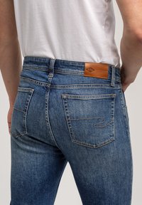 Granatowe dżinsy o gładkiej fakturze, z skórzaną łatą z tyłu, dwoma tylnymi kieszeniami oraz kontrastującym przeszyciem.