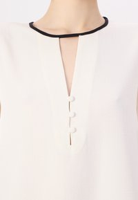 Top in tessuto bianco con scollatura nera, apertura a chiave e tre bottoni decorativi lungo il davanti.