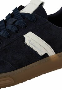 Gros plan d'une basket en daim bleu foncé avec des lacets bleu marine, une bande latérale blanche brandée "tamaris," et une semelle en caoutchouc brun clair.