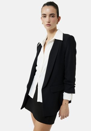 Femme portant un blazer noir sur une chemise blanche à boutons et une jupe noire, avec des boucles d'oreilles anneaux dorées et les cheveux attachés.
