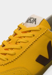 Κίτρινα sneakers από δέρμα, με υφή στην επιφάνεια, αντίθετο καφέ λογότυπο και κίτρινες κορδόνια με ετικέτα υπογραφής στη γλώσσα.