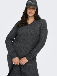 Ung kvinna i mörkgrå stickad klänning och keps med texten "Los Angeles", håller i svarta väskremmar och tittar rakt fram med ett neutralt uttryck.