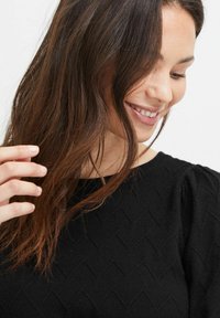 Schwarzer Pullover mit strukturiertem Zickzackmuster, rundem Ausschnitt und schulterpuffigen Ärmeln. Model, das leicht ihr Haar berührt und lächelt.