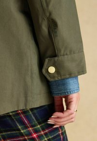 Olivegreenowa rękawiczka z złotym guzikiem, z dżinsowym mankietem pod spodem i widocznym wzorem w kratę w okolicy talii.