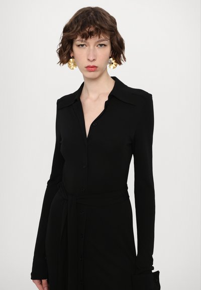 Čierna košeľa s dlhým rukávom, golierovým dizajnom, hladkou tkaninou, so širokým opaskom v páse a veľkými výraznými náušnicami.