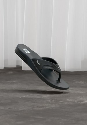 Schwarze Flip-Flop-Sandale der Marke Black Reef schräg in der Luft über einem grauen Betonboden und weißem Vorhang im Hintergrund.