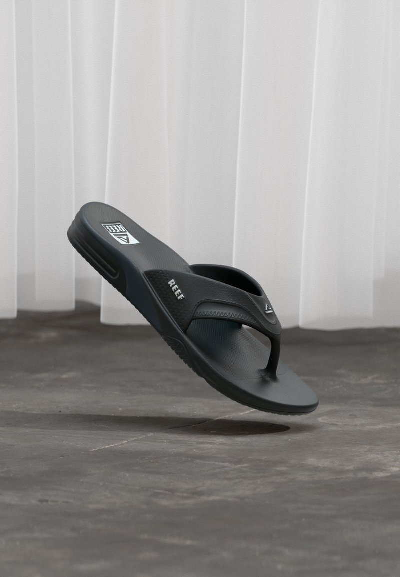 Μαύρο σανδάλι Black Reef τύπου φλιπ-φλοπ σε προσανατολισμό στον αέρα πάνω από γκρι τσιμεντένιο δάπεδο και λευκή κουρτίνα στο φόντο.