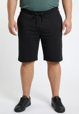 Man draagt zwarte shorts met trekkoord, groen shirt en zwarte sneakers, staand tegen een neutrale achtergrond.