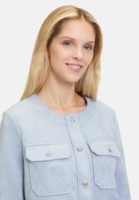 Světle modrá semišová bunda s kulatým výstřihem, stříbrnými knoflíkovými uzávěry a dvěma náprsními kapsami, s texturovanou tkaninou a čistým designem.