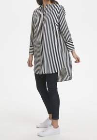 Túnica de rayas en azul marino y crema, con cuello y tapeta abotonada, mangas largas y dobladillo asimétrico. Combinada con vaqueros ajustados oscuros y zapatillas blancas.