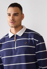Navy blue striped long-sleeve polo shirt with a white collar, featuring a zip closure and a small brown logo on the left chest area.