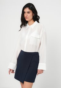 Λευκό, μακρυμάνικο πουκάμισο με τσέπη και κουμπιά στο μπροστινό μέρος, συνδυασμένο με μια μίνι φούστα γραμμή Α σε ναυτικό μπλε, με φερμουάρ στο πλάι. Ομαλές υφές.