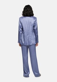 Woman with shoulder-length dark hair wearing a shiny lavender suit with long blazer and wide-leg trousers, standing facing away.