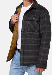 Kurtka w kratę o ciemnoszarym wełnianym wierzchu, pomarańczowym pikowanym podbiciu, dwóch kieszeniach na piersi i czarnych guzikach. Posiada zapięcie na guziki z przodu.