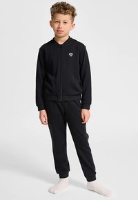 Svart zip-up träningsjacka med en liten logotyp på bröstet, kombinerad med matchande joggingbyxor. Tyget ser mjukt och slätt ut. Modellen står framför en vit bakgrund.