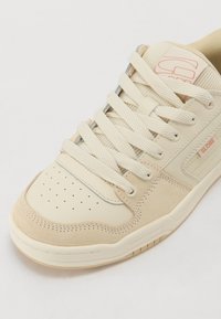 Crème en beige sneaker van textuur suede en glad leer. Heeft een geperforeerde neus, platte veters en subtiele branding aan de zijkant.