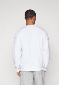 Sweatshirt blanc à manches longues et col rond, avec une texture lisse. Porté sur un pantalon de survêtement gris, montrant l'arrière.