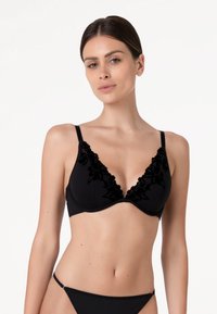 Donna che indossa un reggiseno nero con dettagli in pizzo floreale e slip neri abbinati su uno sfondo bianco semplice.