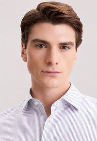 Jonge man met bruin haar, gekleed in een wit shirt met dunne blauwe strepen, kijkt rechtstreeks in de camera tegen een effen achtergrond.