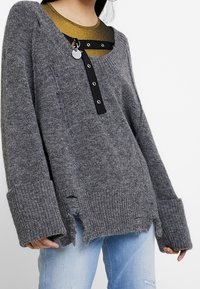 Grau gestrickter Pullover mit V-Ausschnitt, ausgefranstem Saum, Rippenbündchen und schwarzem Knopfverschluss. Über einem metallisch goldenen Oberteil getragen.