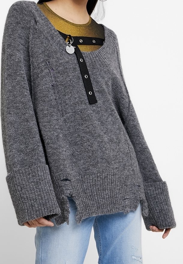 Grau gestrickter Pullover mit V-Ausschnitt, ausgefranstem Saum, Rippenbündchen und schwarzem Knopfverschluss. Über einem metallisch goldenen Oberteil getragen.