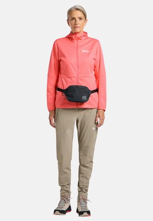 Femme portant une veste zippée corail, un pantalon de randonnée beige, des chaussures de randonnée beiges et un sac banane noir, debout devant un fond clair.