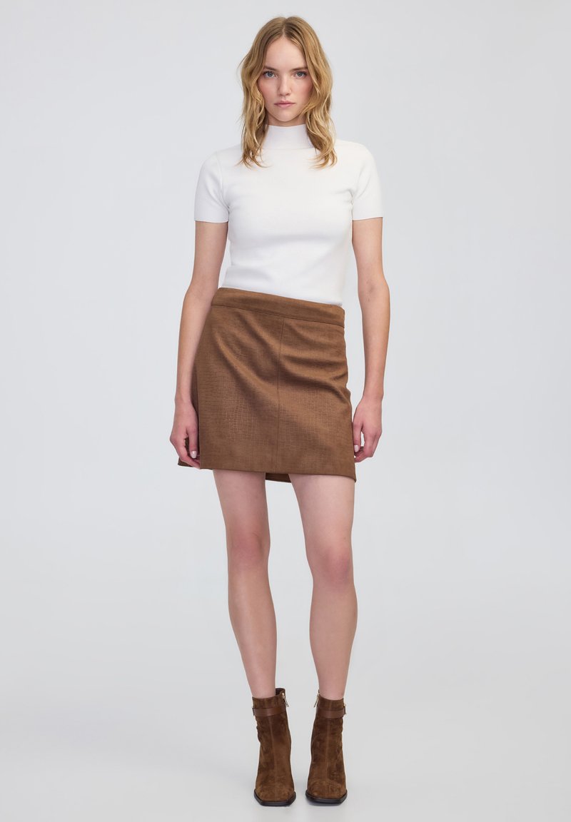 Top blanco de manga corta combinado con una falda mini de cuadros marrones. La modelo lleva botas de tobillo de ante marrón con un tacón grueso.