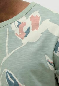 Šviesiai žalias trumpomis rankovėmis marškinėliai su rožių, mėlynų ir baltų gėlių raštu. Minkšta tekstūra, laisvas siluetas ir apvalus iškirpimas.