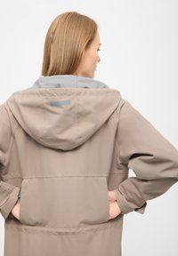 Vrouw van achteren gezien, draagt een beige kapjas met handen in de zakken tegen een effen lichte achtergrond.