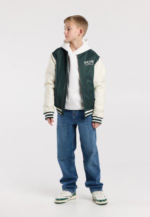 Jongen met blond haar die een groen-wit varsityjack draagt, een witte capuchontrui, blauwe jeans en witte sneakers, en naar links kijkt.