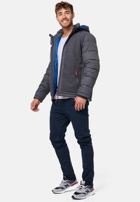 Doudoune grise avec intérieur bleu, capuche à cordon de serrage et deux poches zippées. Associée à un pantalon marine et des baskets grises avec des accents rouges.