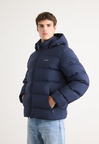 Mornarska puffer jakna s kapuljačom, kviltani dizajn, prednji zatvarač, dva bočna džepa i mali logo na prsima. Mekani, izolacijski materijal.