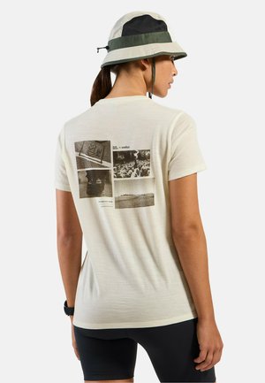 Woman wearing light beige graphic t-shirt and green-and-beige bucket hat, shown from back, with black shorts and wristwatch.