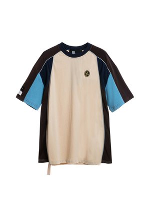 Joven con una camiseta de colores beige, marrón y azul y pantalones cortos negros con ribetes azules y grises, de pie sobre un fondo blanco.