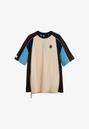 Joven con una camiseta de colores beige, marrón y azul y pantalones cortos negros con ribetes azules y grises, de pie sobre un fondo blanco.