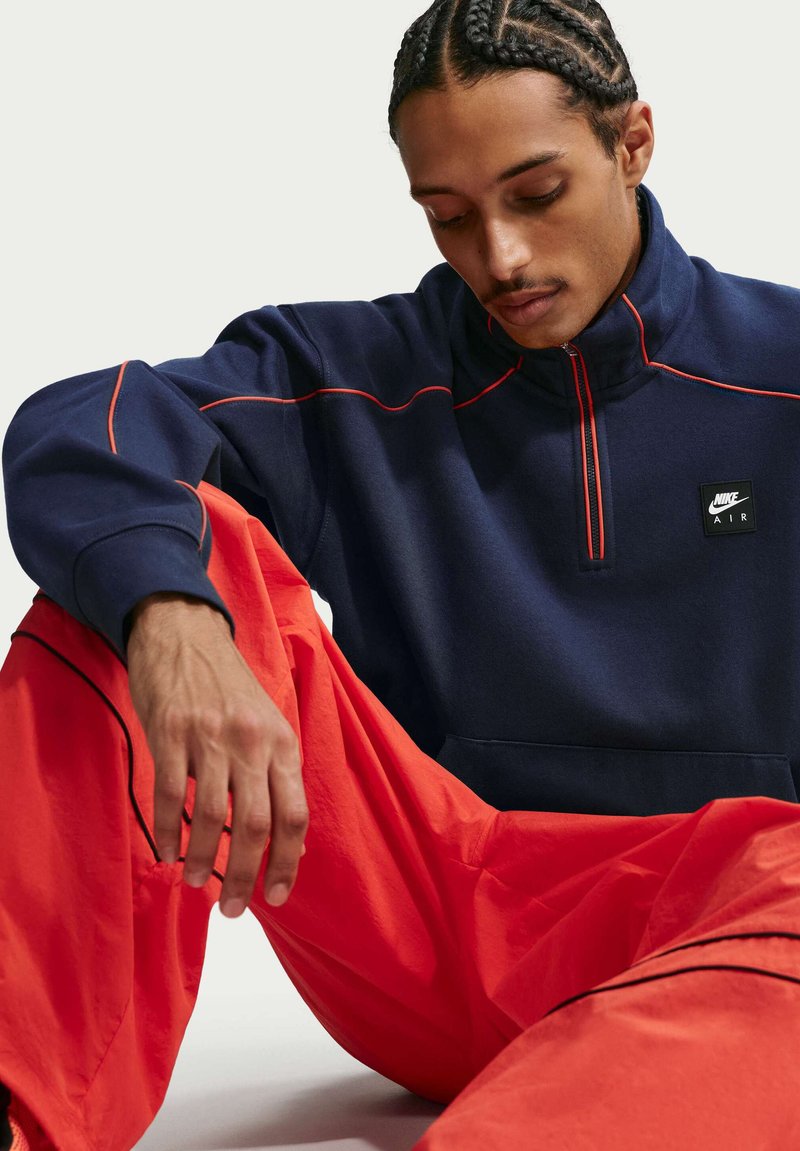 Homme aux cheveux tressés portant un sweat-shirt Nike Air à demi-fermeture éclair bleu marine et un pantalon orange vif, assis les yeux fermés et la tête inclinée vers le bas.