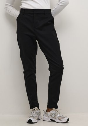 Person trägt schwarze Slim-Fit-Hose und weiße Turnschuhe, steht vor einer schlichten, hellen Wand.